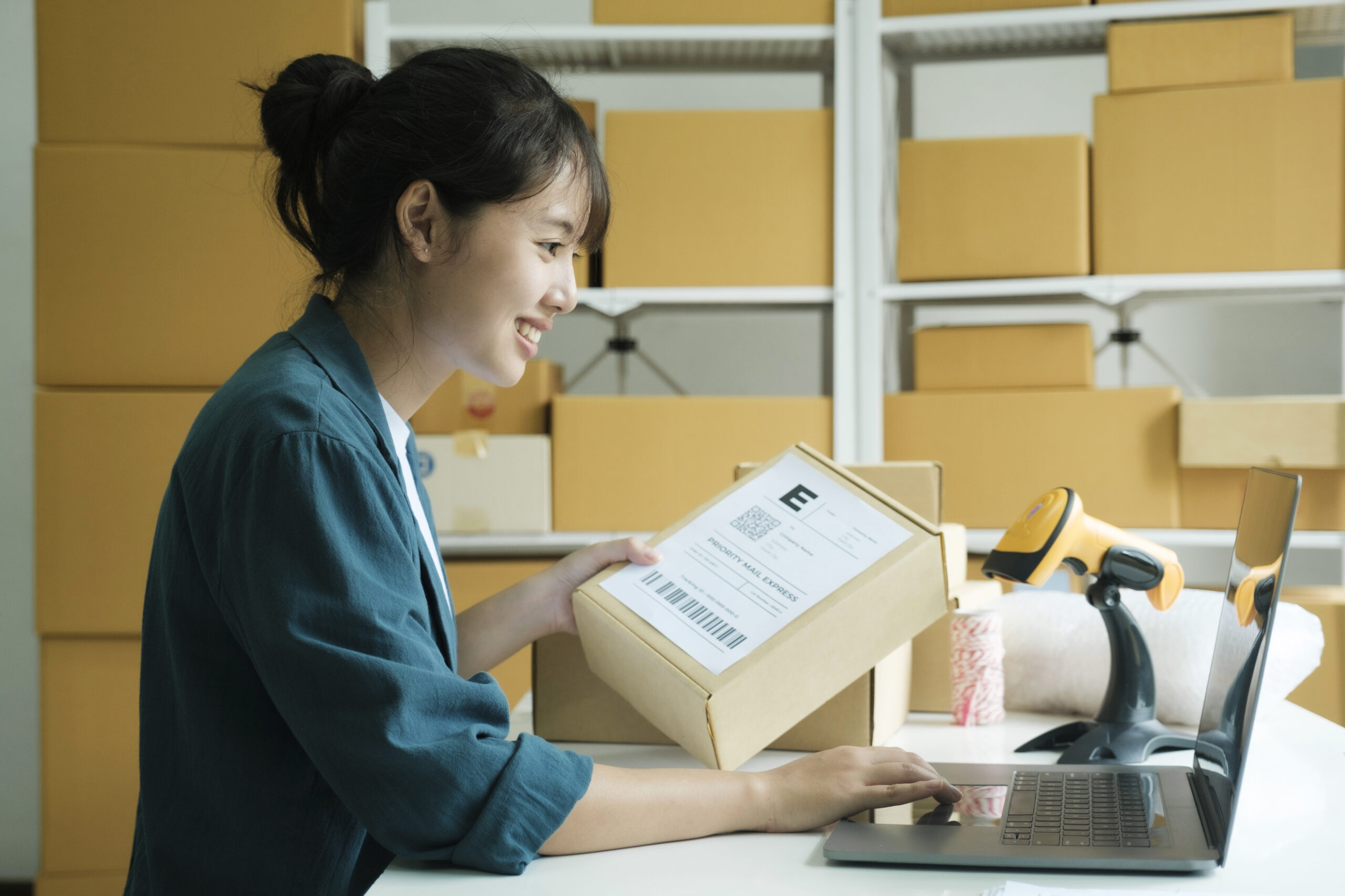 Young female small business entrepreneur, online store owner holding parcel checking address before delivery and shipping to customer using laptop at warehouse, workplace. Online selling, e-commerce conept.